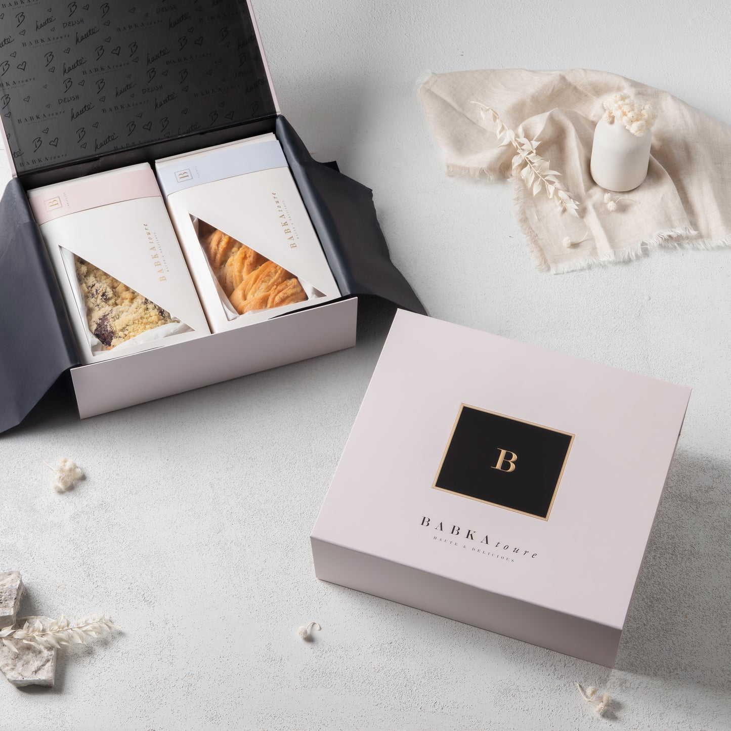 A pale pink Babkatoure Large Signature Edition Gift Box 1 sits on a white surface. Next to it, an open box reveals two delicious babkas—Chocolate and Vanilla—surrounded by a candle, decorative cloth, and flowers.