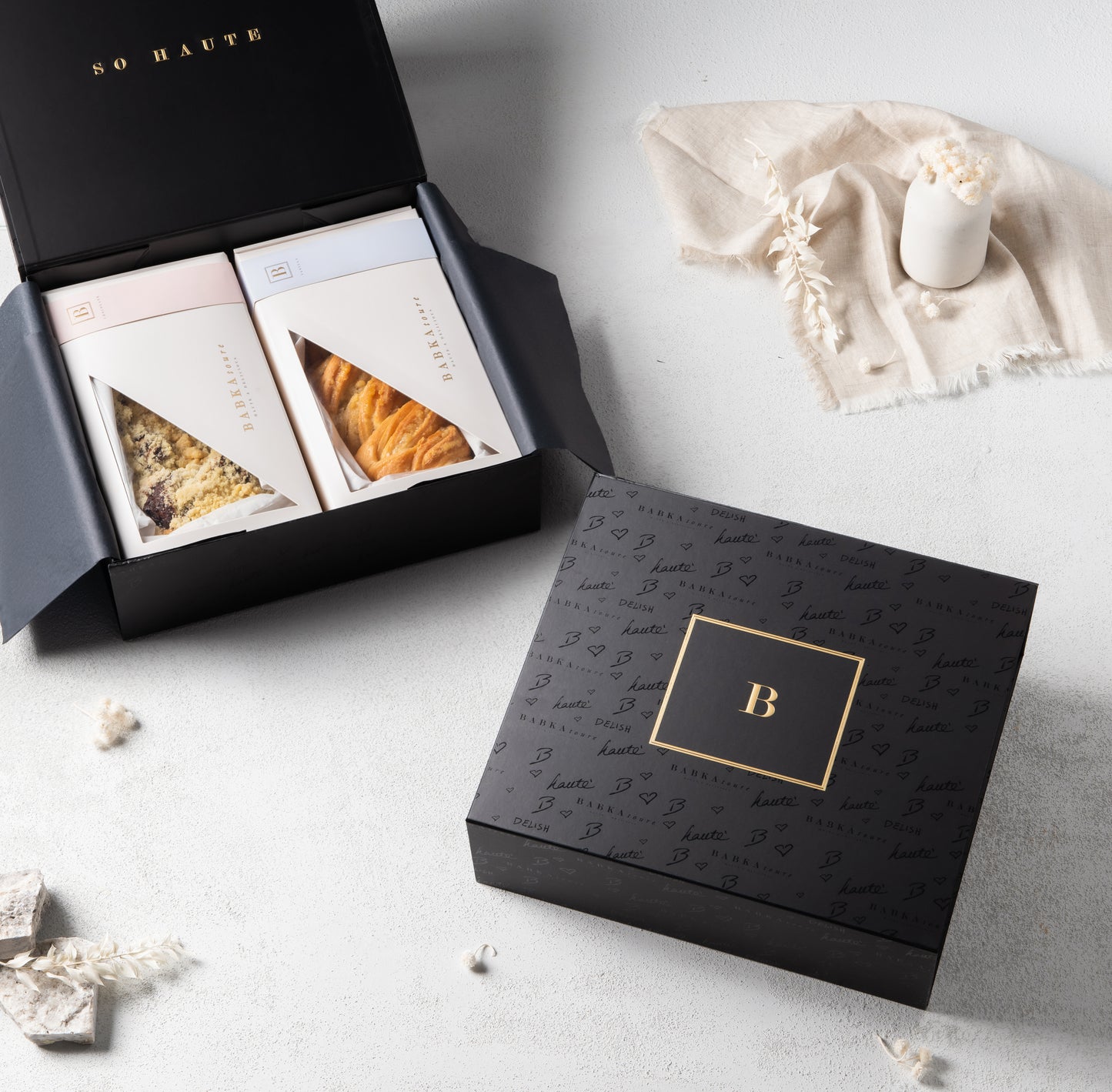 An open Large Midnight Edition Gift Box 1 by Babkatoure, featuring a black box with a gold B logo and two packaged chocolate babka pastries inside, sits beside a closed matching box on a light surface with candle and decorative accents.