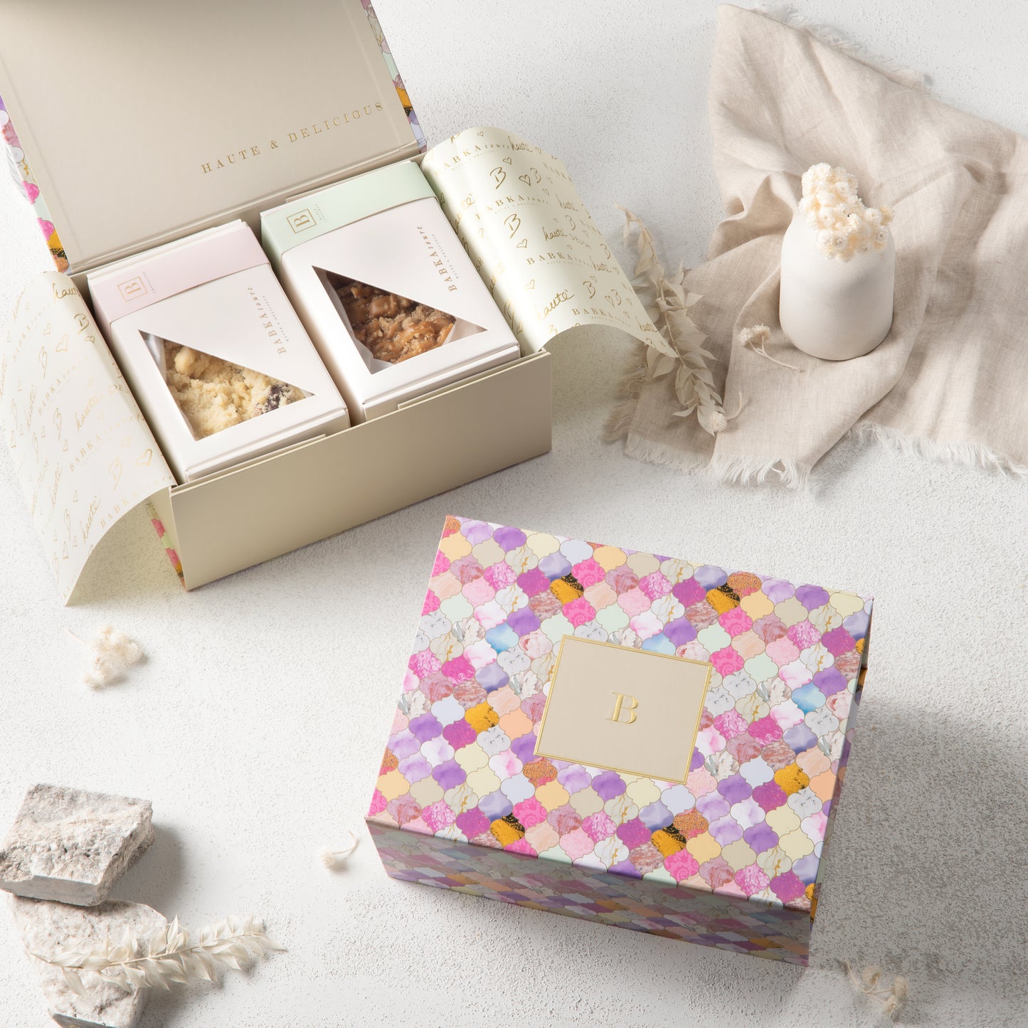 The Babkatoure Medium Mosaic Edition Gift Box features a pastel box with a gold B beside an open box displaying three snacks, including mosaic-textured white treats, styled with a white candle and dried flowers on a light surface.