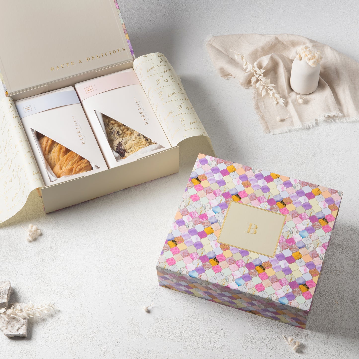 The Babkatoure Large Mosaic Edition Gift Box 1, featuring a gold B on the lid, is shown beside an open box filled with chocolate and vanilla pastries, all arranged on a light surface with dried flowers and a candle.
