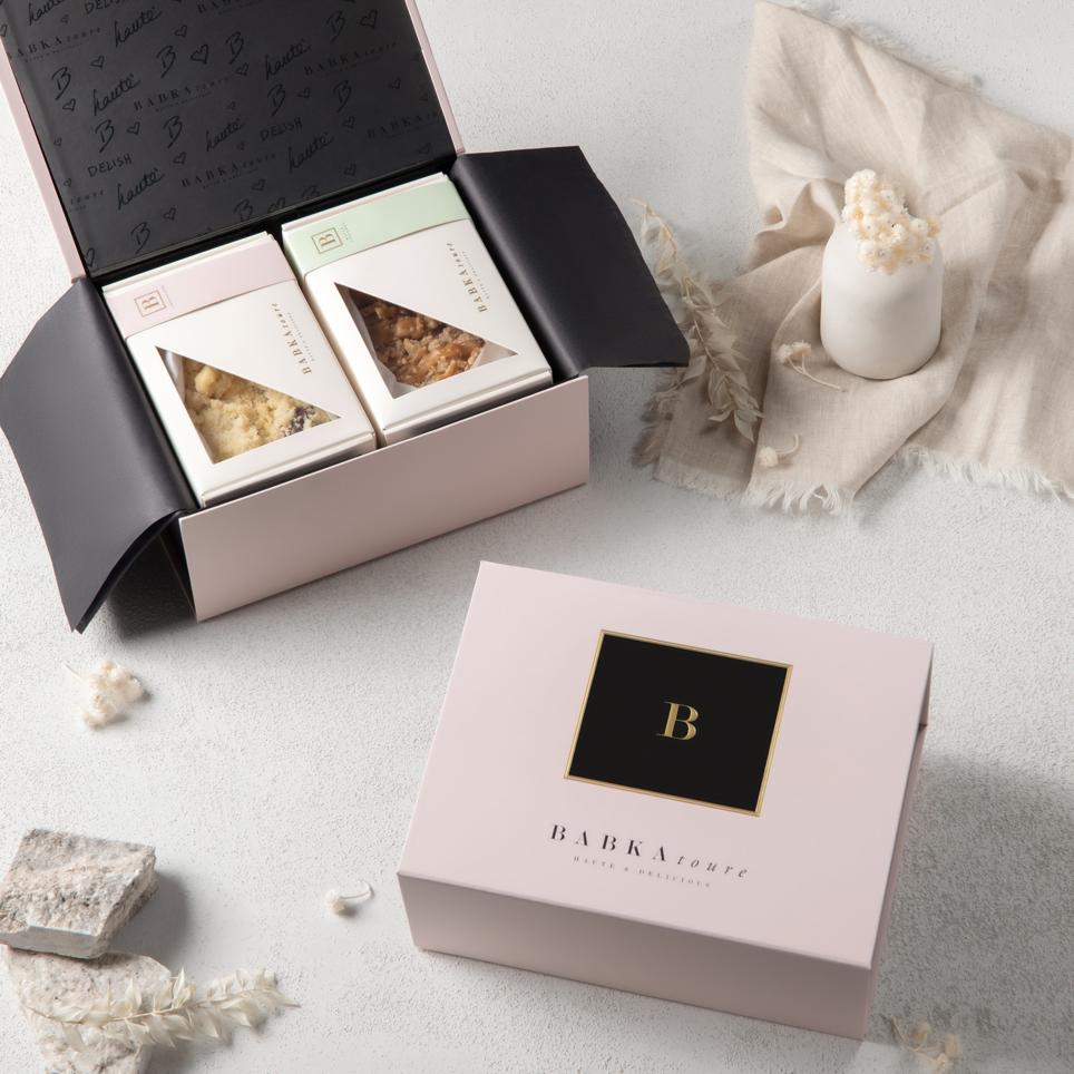 A pale pink Babkatoure Medium Signature Edition Gift Box labeled BABKA holds two individually boxed pastries. The open box rests on a light surface with a candle, fabric, and stones nearby in an elegant, minimal display.