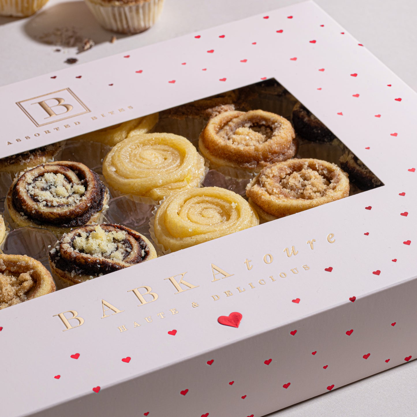 The Babkatoure Amour Edition 24pc Babka Bites Box (Assorted) features a white box with red hearts and gold lettering, displaying swirled Cinnamon and Chocolate Bites through a clear window.